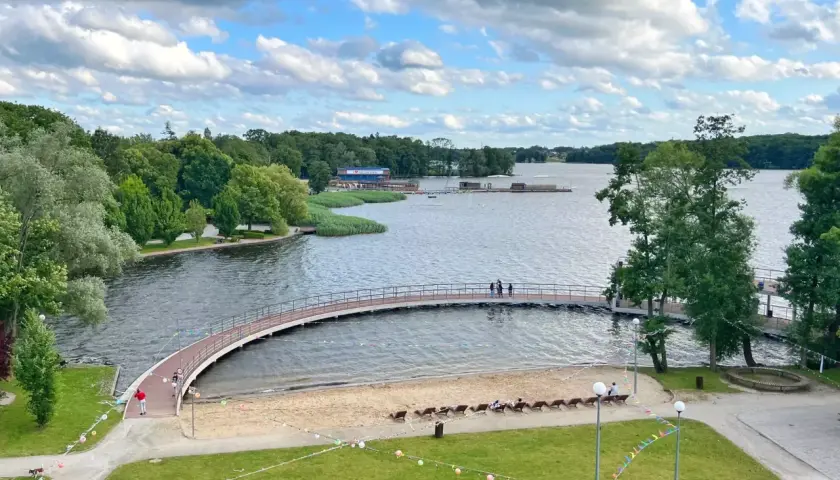 Malownicze jezioro w Szczecinku z łukowatym mostkiem, piaszczystą plażą i zielonymi terenami rekreacyjnymi.