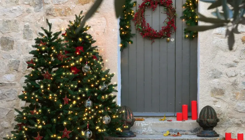 Árbol de Navidad iluminado y una corona de acebo adornan la entrada de una casa de piedra.