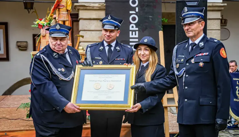 Członkowie OSP otrzymują odznaczenie. W tle drewniana figura i baner.