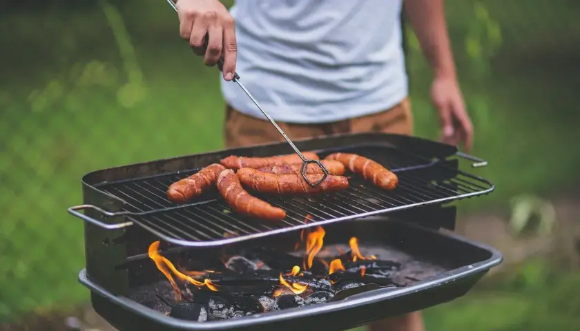 Un homme retourne des saucisses sur un barbecue bois ou charbon. Les flammes dansent sous la grille, promettant un repas savoureux.