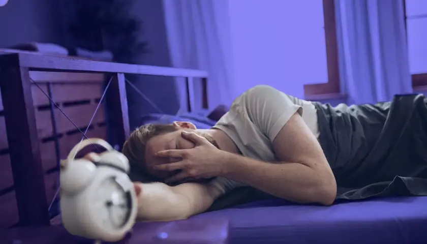 Hombre en la cama, con la cara entre las manos, junto a un despertador. Parece que me levanto cansado.
