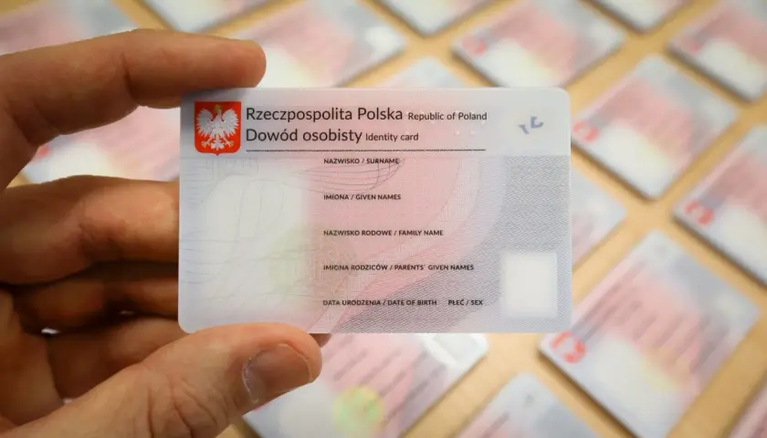 Dłoń trzyma polski dowód osobisty, jeden z wielu leżących na stole. To ważny dokument, zawierający Twój osobisty numer identyfikacyjny.