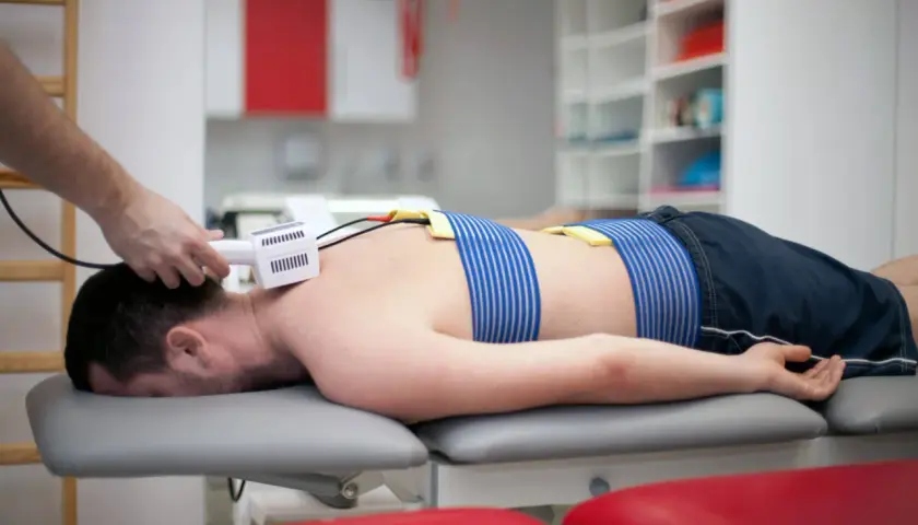 Pacjent leży na kozetce, poddawany zabiegowi z użyciem prądów Traberta. Elektrody przymocowane są do jego pleców.