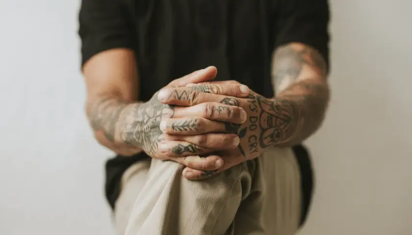 Dłonie z tatuażem handpoke, ozdobione wzorami i napisem "Boys Do Cry", splecione na kolanach.
