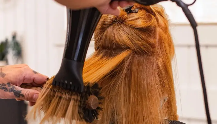 Manos con tatuajes secan y peinan cabello pelirrojo con secador y cepillo redondo. El brushing que es, es una técnica de estilismo.