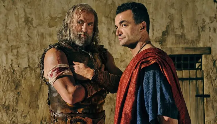 Två män i antika kläder, en med skägg och läderrustning, den andra i röd mantel, står nära varandra. Kanske en scen från Spartacus: House of Ashur.