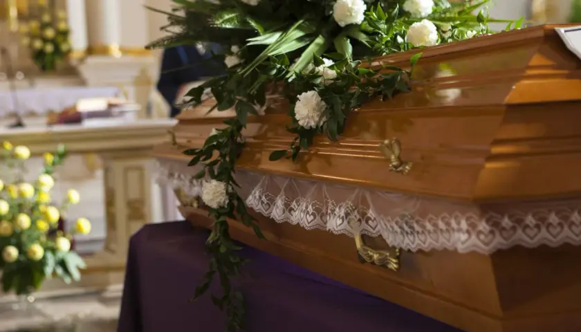 Drewniana trumna ozdobiona kwiatami i koronką, przygotowana do pogrzebu. W tle kościół.