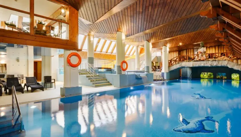 Basen w hotelu Belvedere Resort & Spa z drewnianym sufitem i mostkiem. Woda jest błękitna, a na dnie widoczne są ozdobne ryby.