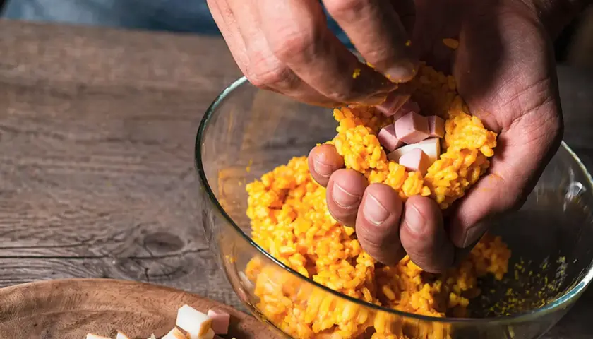 Mani che preparano polpette di riso con cubetti di prosciutto e formaggio.