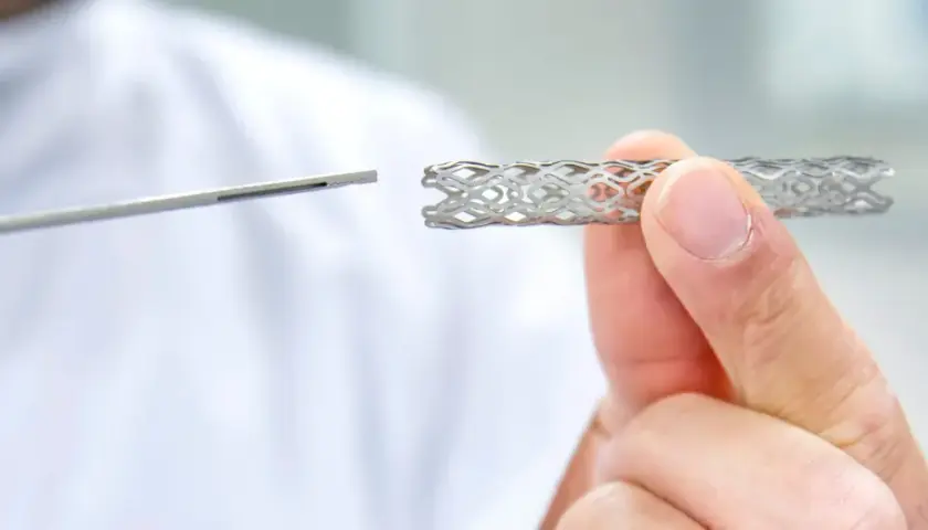 Dłoń trzyma metalowy stent o ażurowej strukturze, obok widoczny jest instrument medyczny.