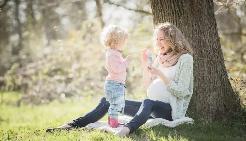 Ciężarna kobieta i małe dziecko puszczają bańki mydlane pod drzewem. Mama uśmiecha się, wiedząc, że robi wszystko, żeby cię chronić.
