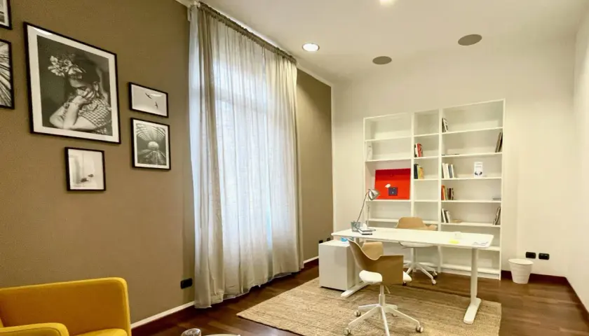 Arredamento studio psicologo: scrivania bianca, libreria, sedie, poltrona gialla, quadri e tenda.