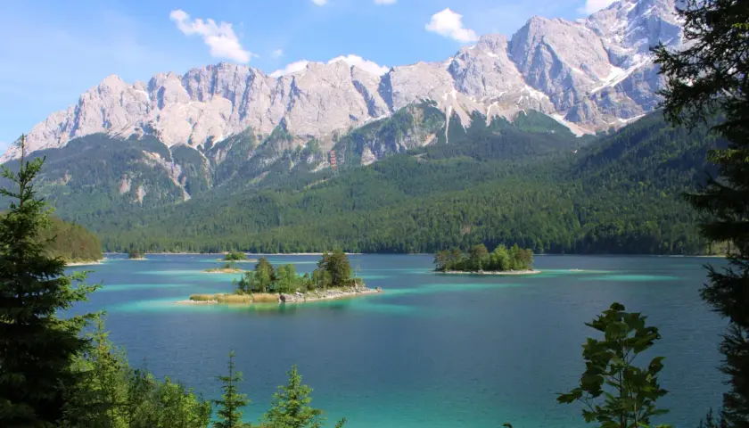 Turquoise lake with islands dotted with trees, framed by a dense green forest and majestic, snow-capped mountains. A quintessential view of the Bavarian countryside.