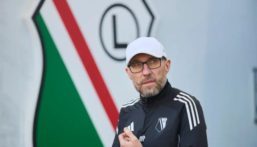 Nowy trener Legii w białej czapce i okularach, na tle klubowego herbu.