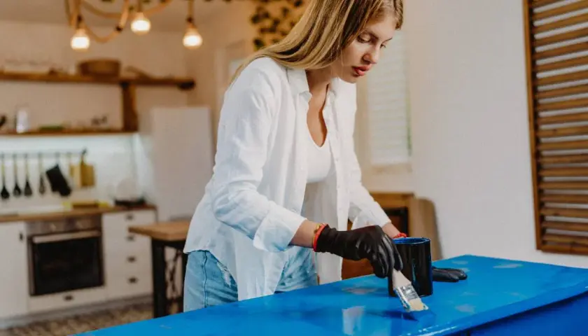 Donna con guanti neri applica vernice blu su un mobile, trasformandolo per rendere opaco un mobile lucido.