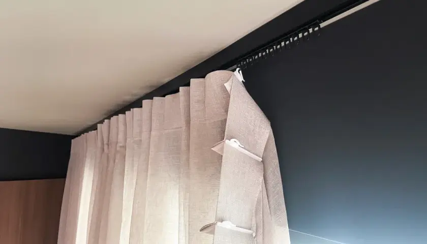 Jasne, lniane zasłony z białymi klipsami, zawieszone na czarnej szynie sufitowej. Eleganckie fleksy do firan zapewniają idealne ułożenie materiału.