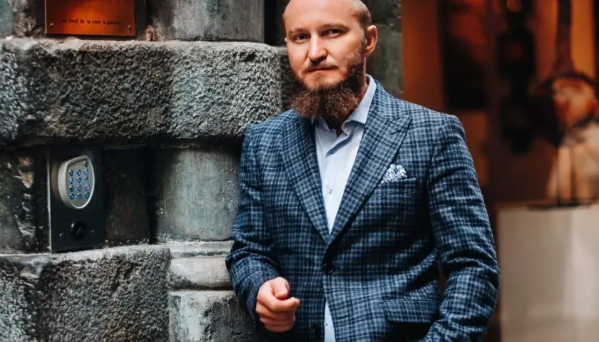 Elegancki mężczyzna w garniturze w kratę, z brodą, oparty o kamienną ścianę. Idealny przykład, jaki krawat do garnituru w kratę wybrać.