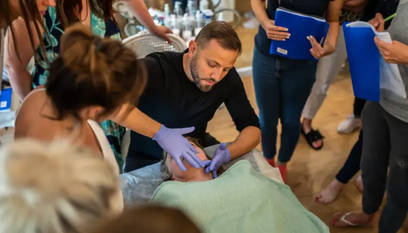 Masaż Kobido co to? Szkolenie z liftingującego masażu twarzy. Terapeuta w rękawiczkach demonstruje techniki na leżącej osobie, obserwowanej przez grupę.