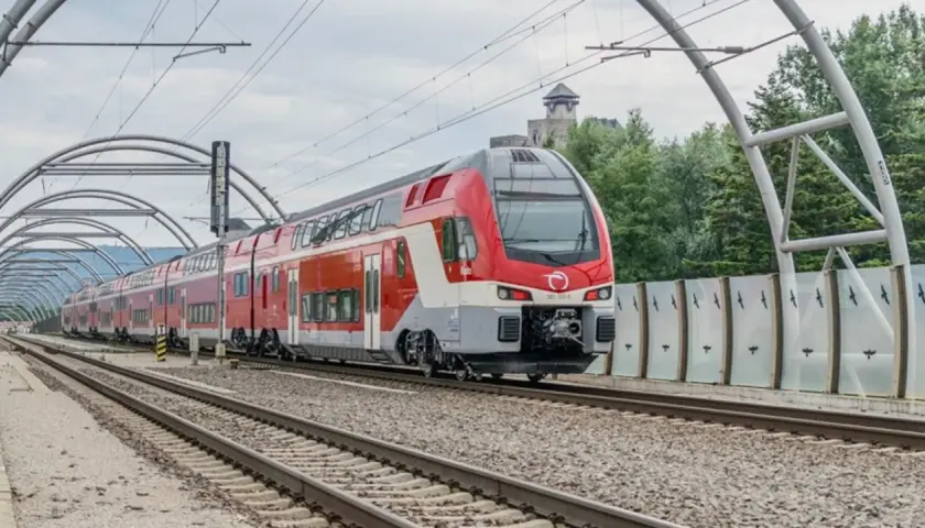Moderner Doppelstockzug der **Slovak Railways** fährt unter einer Bogenkonstruktion auf Gleisen. Im Hintergrund eine Burg.