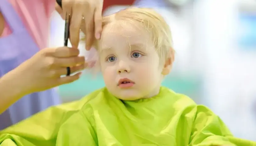 Mały chłopiec z blond włosami przeżywa swoje pierwsze strzyżenie dziecka. Fryzjerka delikatnie podcina mu włosy.