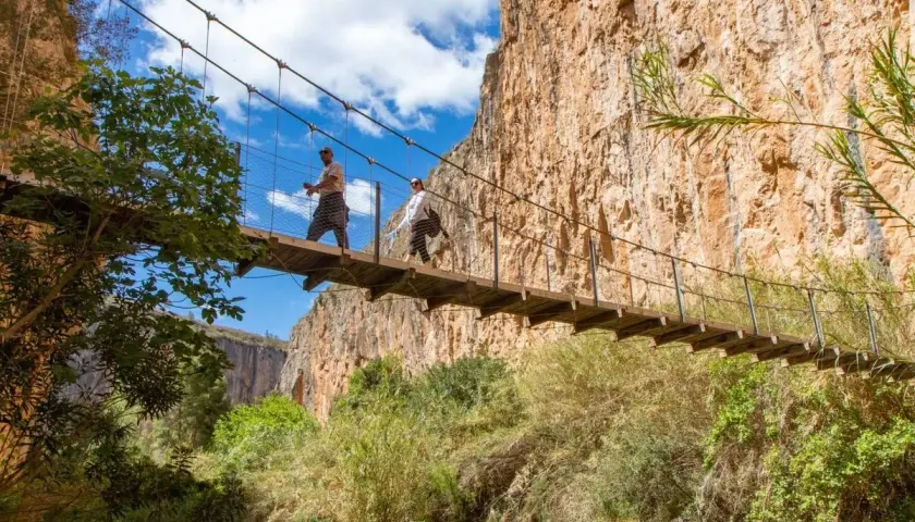 Dos excursionistas cruzan un puente colgante sobre un río, explorando las rutas de senderismo de Valencia.