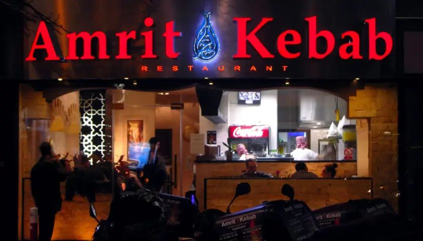 Jasny neon "Amrit Kebab" nad wejściem do restauracji. W tle widać wnętrze z pracującymi kucharzami i lodówką Coca-Coli. Na pierwszym planie stoją skutery z pudełkami na dowóz.