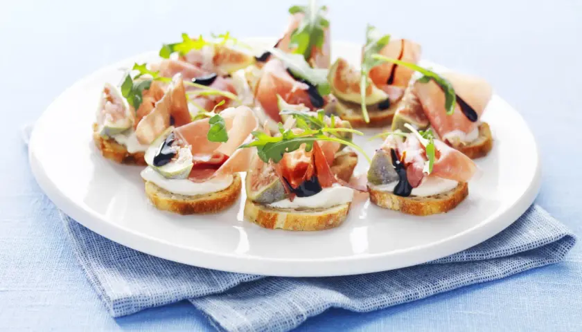 Köstliche italienische Crostini mit Prosciutto, Feigen, Mozzarella und Rucola auf einem weißen Teller.