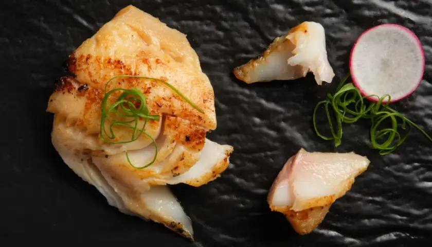 A beautifully grilled saikyo yaki fish fillet, garnished with green onions and a radish slice, rests on a black plate.