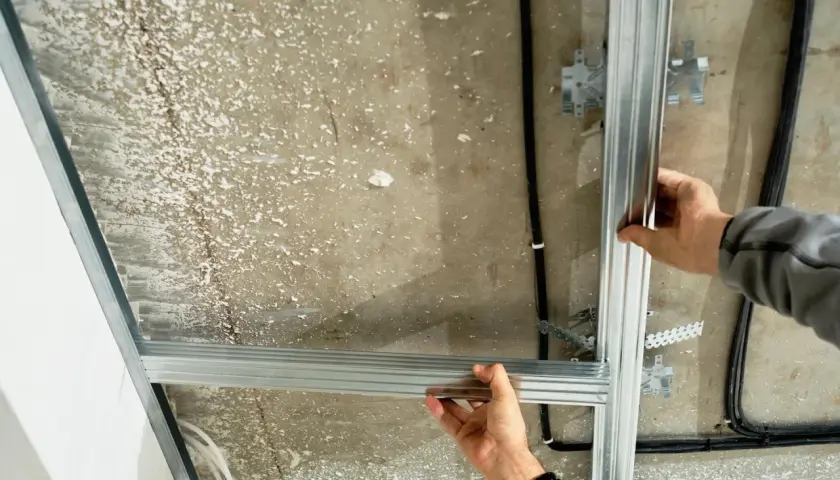 Ręce montują konstrukcję sufitu podwieszanego, łącząc profile aluminiowe. Widać kable i elementy mocujące.