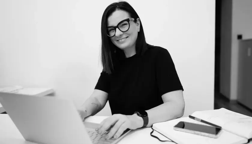 Kobieta w okularach pracuje na laptopie przy biurku z notatnikiem i telefonem.