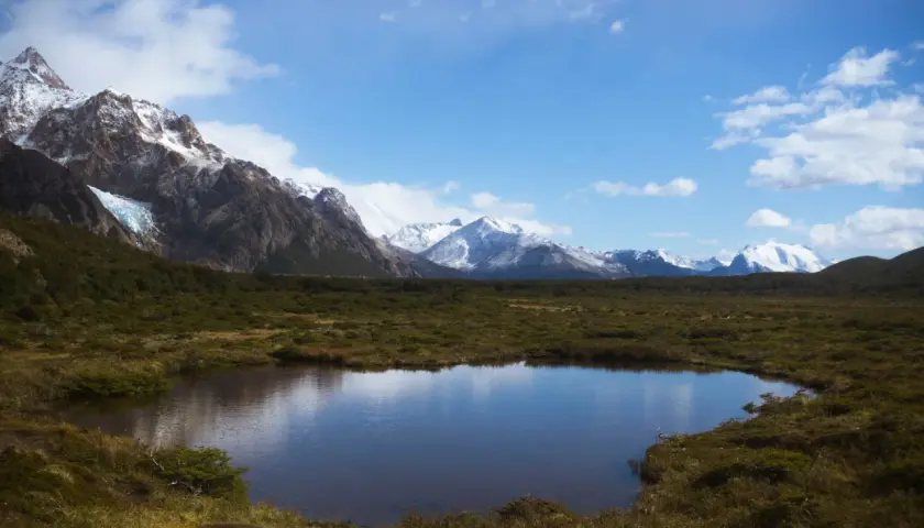 Spokojne jezioro odbija ośnieżone góry, w tym majestatyczne góry z Aconcagua, otoczone zielonymi wzgórzami pod błękitnym niebem.