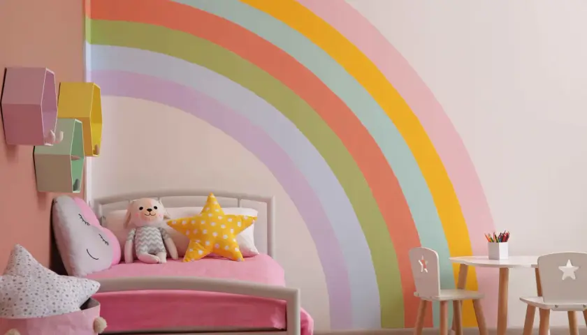 Pokój dziecięcy z pastelową tęczą na ścianie, łóżkiem i stolikiem. Inspiracja, jak namalować tęczę na ścianie.