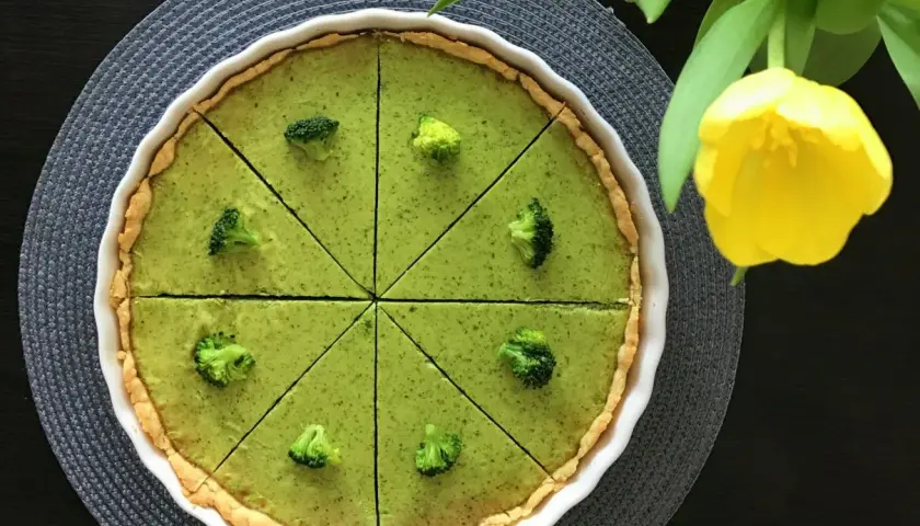 Zielona tarta z brokułami, pokrojona na 8 kawałków, ozdobiona różyczkami brokułów. Obok żółty tulipan.