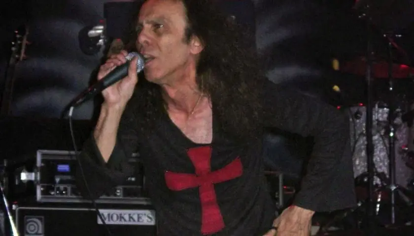 Ronnie James Dio zespół śpiewa do mikrofonu, ubrany w czarną koszulkę z czerwonym krzyżem.