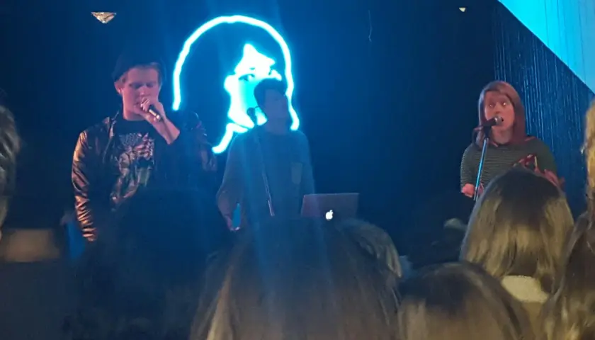 Koncert zespołu tv girl. Na scenie trójka artystów, w tle neonowa podobizna twarzy.