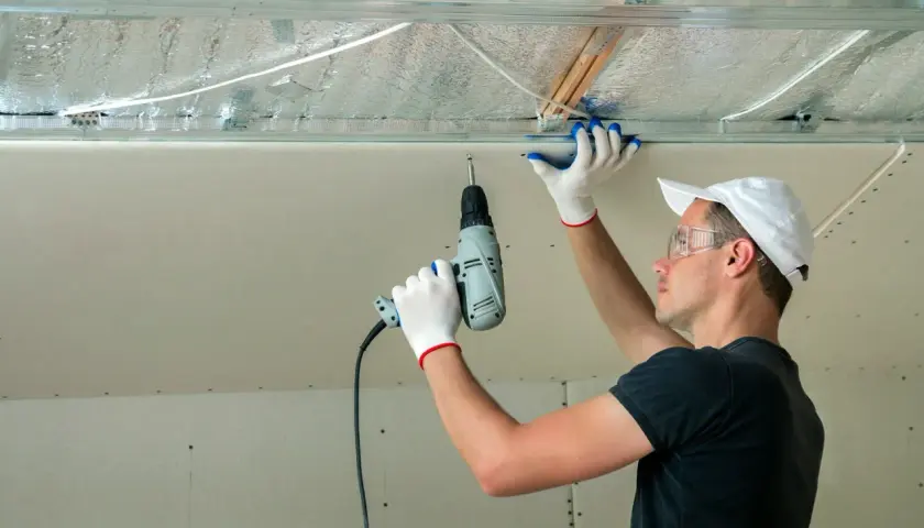 Un ouvrier installe des isolants minces sous un faux plafond en plaques de plâtre, utilisant une perceuse pour fixer les éléments.