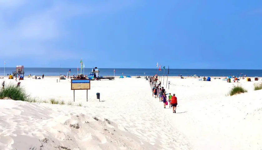 Menschen spazieren auf einem Holzsteg über den weiten, weißen nordsee strand. Der Himmel ist blau, das Meer ruhig.