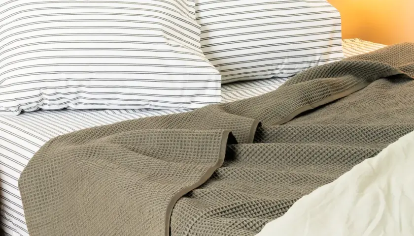 A textured, grey blanket is draped at the end of a bed, over striped pillows and sheets.