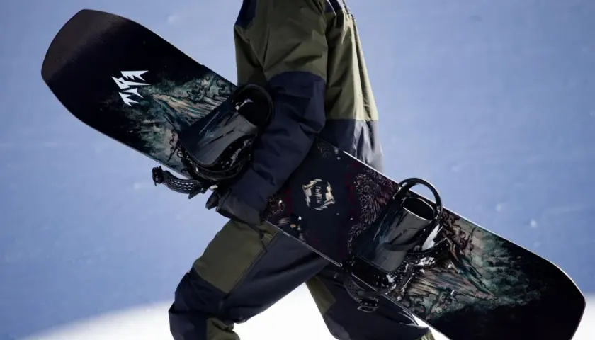 Osoba w zimowym stroju niesie dwa snowboardy z grafiką. Jeden z nich ma logo "JONES".
