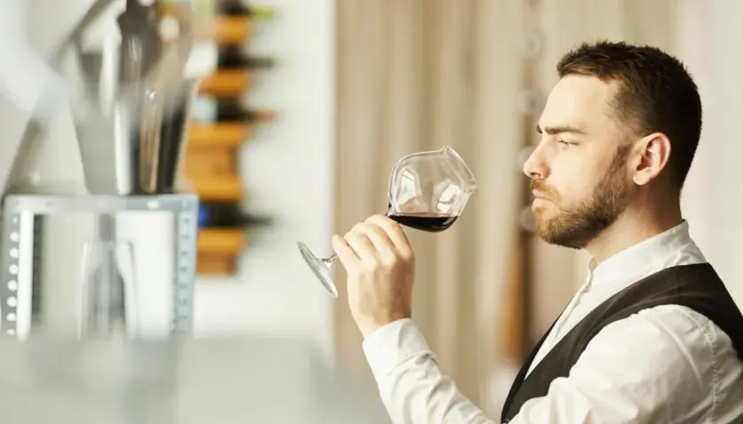 Mężczyzna w białej koszuli i kamizelce, sommelier, kto to? Wącha kieliszek czerwonego wina, oceniając jego aromat.