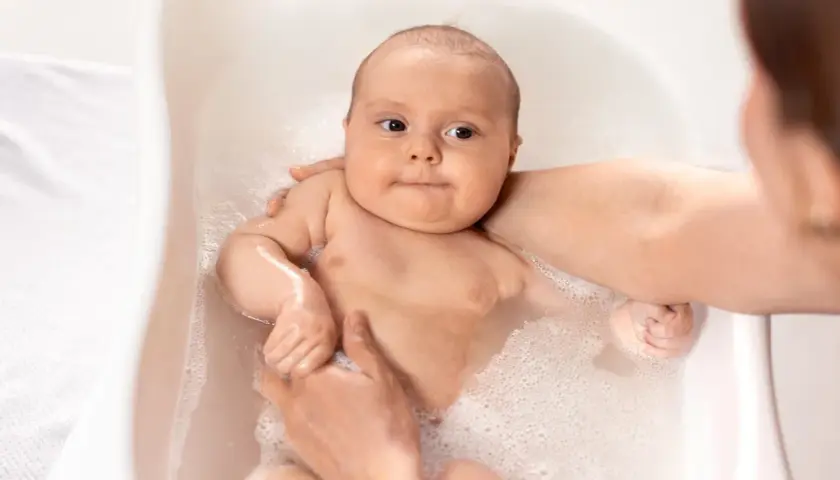 Söt bebis njuter av sitt bad. Händerna håller försiktigt i den lilla kroppen medan bubblorna dansar runt.