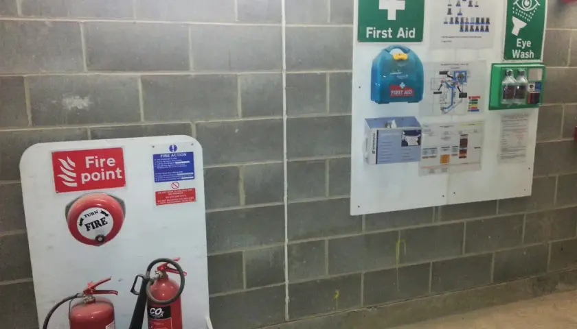 Na ścianie z bloczków betonowych widoczne są znaki budowlane: punkt alarmowy z gaśnicami, apteczka pierwszej pomocy i instrukcje bezpieczeństwa.