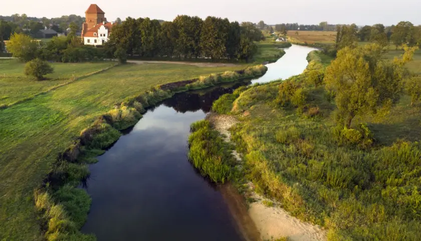 Malownicza wieś nad Liwcem, z charakterystycznym dworem i wieżą, otoczona zielonymi łąkami i drzewami. Rzeka Liwiec meandruje przez krajobraz.