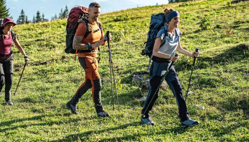 Trzy osoby wędrują po zielonym zboczu, ubrane w wygodne, techniczne spodnie w góry i z plecakami.