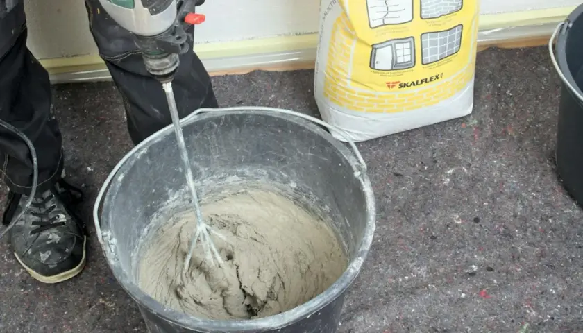 Personen använder en borrmaskin för att blanda betong i en hink. En gul säck med Skalflex-märkning står bredvid.