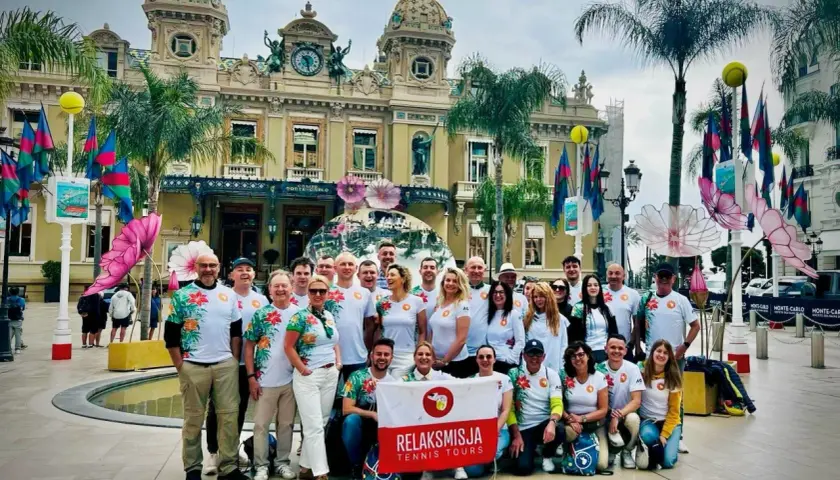 Grupa turystów z flagą "RelaksMisja Tennis Tours" przed kasynem w Monte Carlo. Wszyscy ubrani w koszulki z motywem kwiatowym.