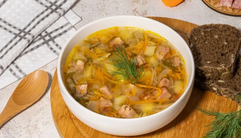 Aromaticzny kapuśniak ze słodkiej kapusty z kawałkami mięsa, ziemniakami i marchewką, udekorowany koperkiem. Obok kromki ciemnego chleba.