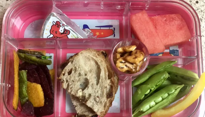 A pink bento box with watermelon, peas, pretzels, bread, and veggie sticks. Add an ice pack to keep this bento box cold.
