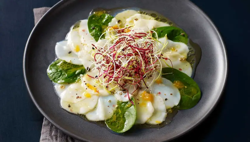 Carpaccio de Saint-Jacques aux agrumes, garni de pousses croquantes et de feuilles d'épinard frais, arrosé d'une vinaigrette légère.