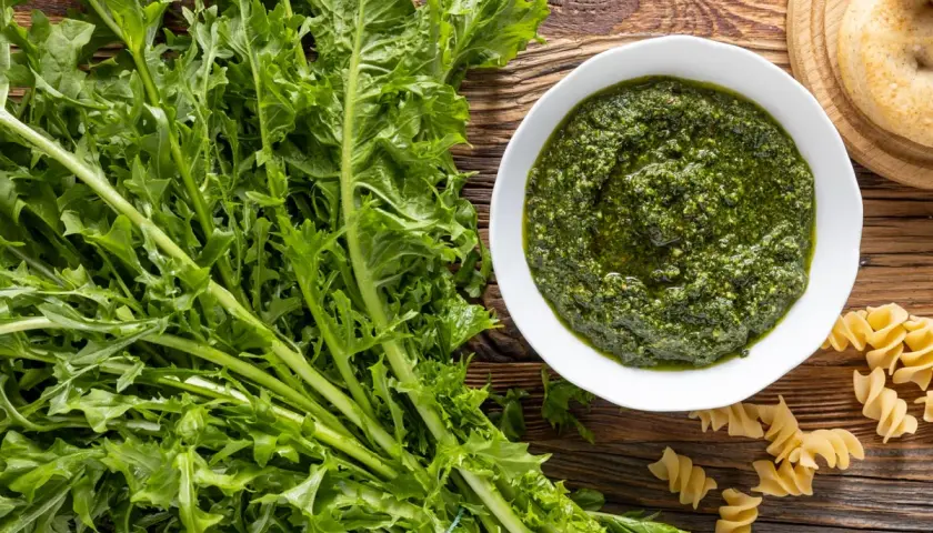 Un mazzo di **tarassaco foglie** fresche accanto a una ciotola di pesto verde brillante e pasta fusilli su un tavolo di legno rustico.